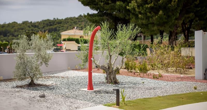 Doccia solare da giardino installata in un'area verde con pavimentazione drenante e ulivi ornamentali.
